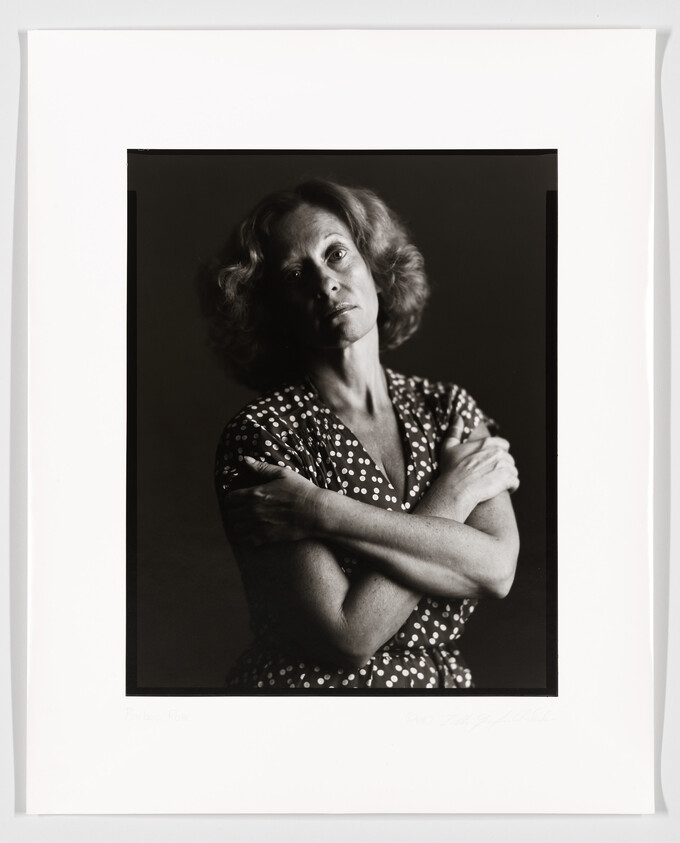 Black and white portrait of a woman with curly hair, wearing a polka dot dress, arms crossed, looking pensively to the side. The photograph is framed with a white border, and there is a signature at the bottom.