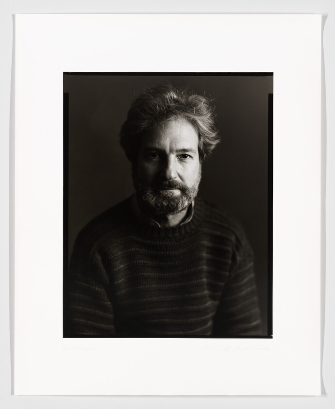 A black and white portrait of a man with a thoughtful expression, featuring tousled hair and a beard, wearing a dark striped sweater. The photo is framed with a white border that includes a signature and text at the bottom.