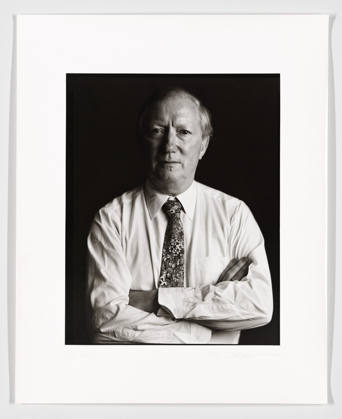 Black and white portrait of a middle-aged man with fair skin, wearing a white shirt and patterned tie, sitting with his arms crossed. The photograph is framed with a white border and has a signature at the bottom right corner.