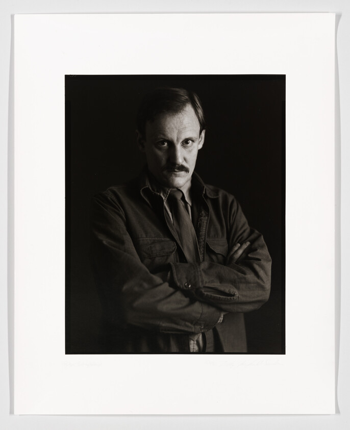 Black and white portrait of a mustached man with his arms crossed, wearing a buttoned shirt and jacket, looking directly at the camera. The photo is framed with a white border, and there is a signature at the bottom right corner.