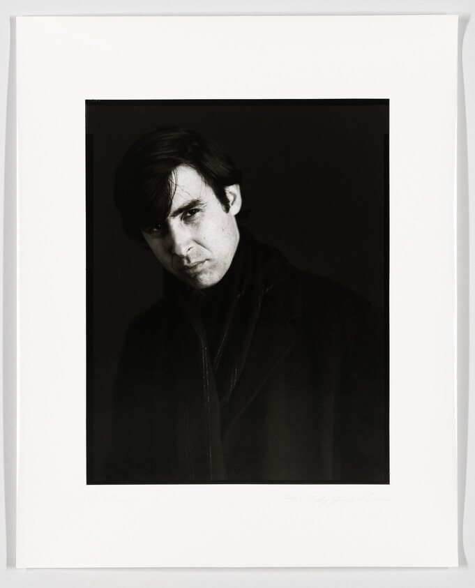 Black and white portrait of a man with dark hair and a serious expression, wearing a dark coat. The photo is framed with a white border that includes a signature and text at the bottom.