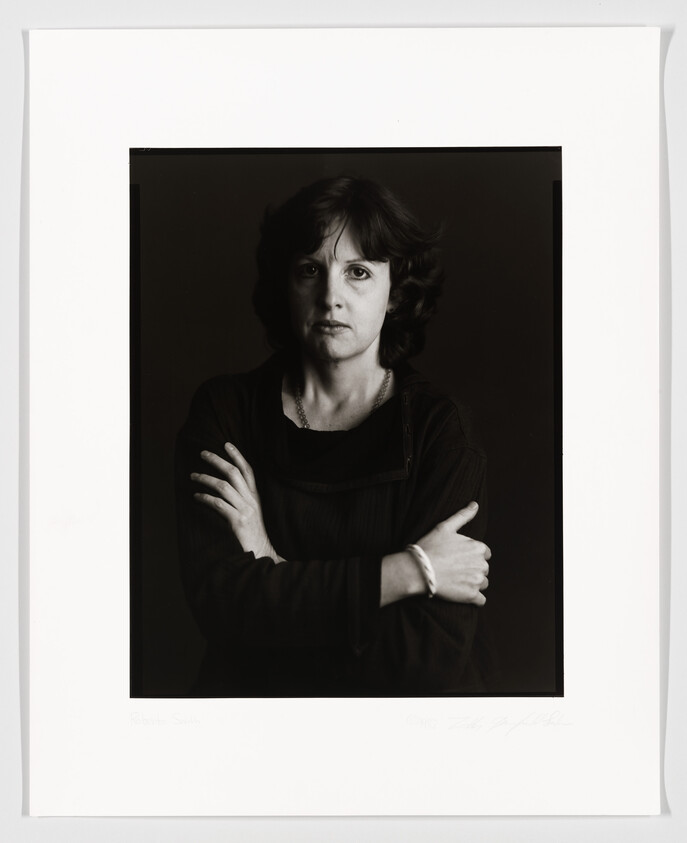 A black and white portrait of a woman with shoulder-length hair, looking directly at the camera with a neutral expression. She is wearing a dark sweater and has her arms crossed. The photo includes a white border with a signature and text at the bottom.