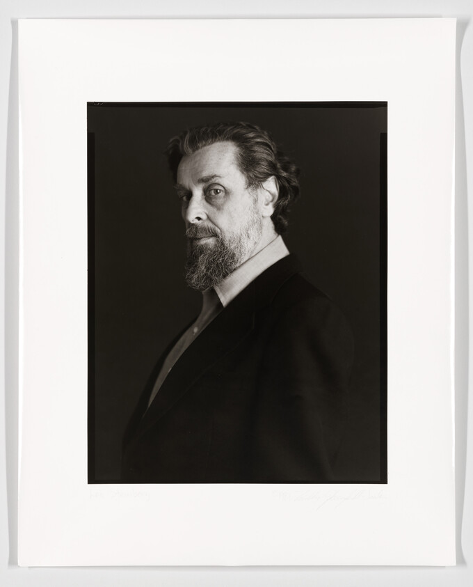 A black and white portrait of a bearded man in a dark suit, looking to the side with a serious expression. The photograph is mounted on white paper with a signature and text at the bottom.