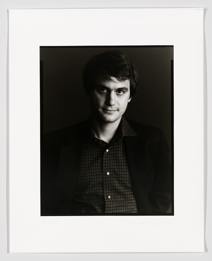 Black and white portrait of a man with dark hair, wearing a checkered shirt and a jacket, looking directly at the camera with a subtle smile. The photo is framed with a white border that includes a signature at the bottom.