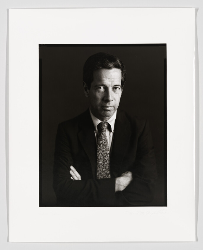 A black and white portrait of a man in a dark suit with a patterned tie. He has his arms crossed and is looking directly at the camera with a serious expression. The photograph is framed with a white border, and there is a signature at the bottom.