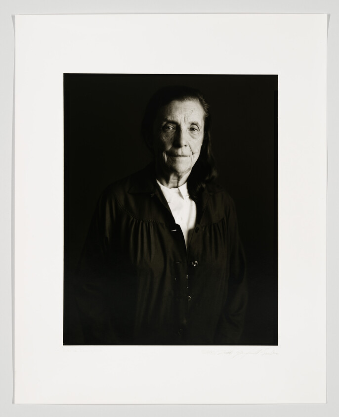 A black and white portrait of an elderly woman with a calm expression, wearing a dark buttoned coat over a light shirt. The photo is framed with a wide white border, and there is a signature at the bottom right corner.