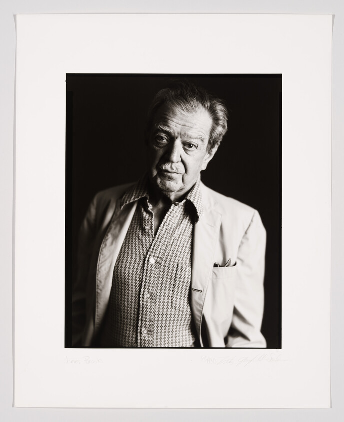 A black and white portrait of an elderly man with a stern expression, wearing a patterned shirt and a light jacket. The photo is framed with a white border that includes a signature and a name at the bottom.