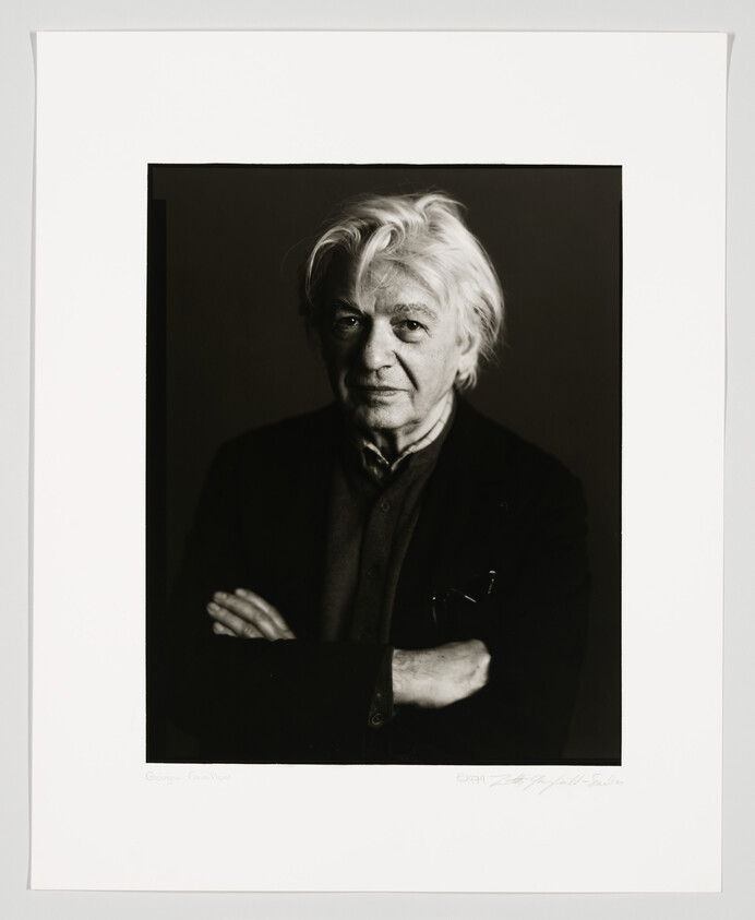 Black and white portrait of an elderly man with white hair, looking directly at the camera with a subtle expression. He is wearing a dark jacket over a collared shirt and has his arms crossed. The photo is framed with a white border, and there are signatures at the bottom.
