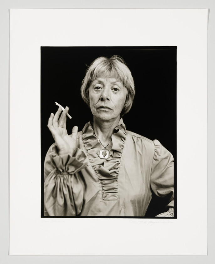 Black and white portrait of a woman with short hair, wearing a blouse and a necklace, holding a cigarette between her fingers. The photograph is framed with a white border, and there is a signature at the bottom right corner.