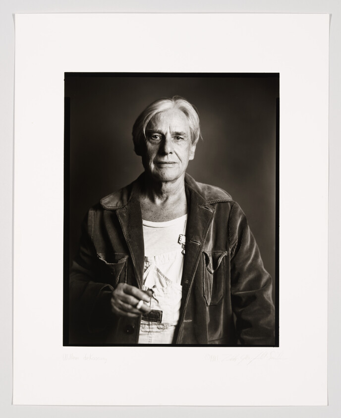 A black and white portrait of an older individual with shoulder-length hair, wearing a jacket over a light shirt, holding a cigarette. The photo is framed with a white border that includes a handwritten signature and text at the bottom.
