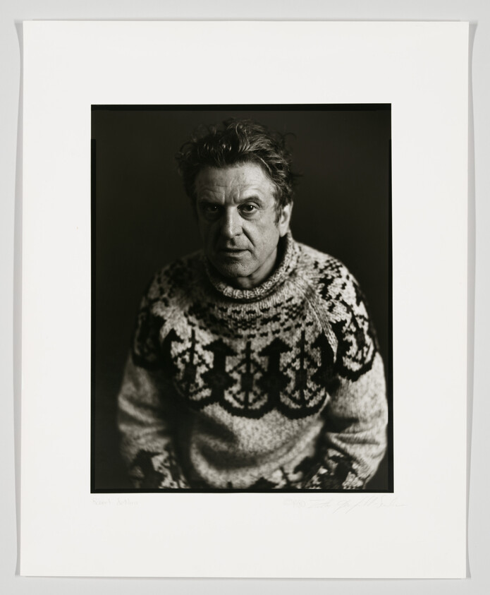 Black and white portrait of a person with tousled hair wearing a patterned sweater, looking directly at the camera with a serious expression. The image is framed with a white border that includes a signature and text at the bottom.