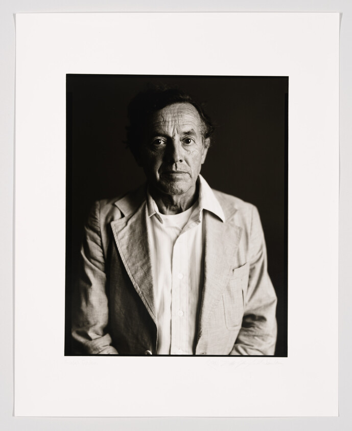 A black and white portrait of an elderly man with expressive eyes and prominent facial lines, wearing a light-colored jacket over an unbuttoned shirt. The photo is framed with a white border that includes a signature at the bottom.