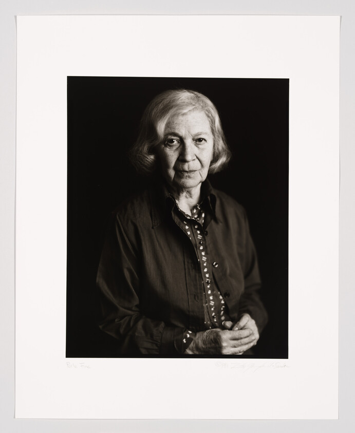 A black and white portrait of an elderly woman with shoulder-length hair, wearing a dark shirt and patterned blouse. She is looking directly at the camera with a neutral expression. The photograph is framed with a wide white border, and there are signatures at the bottom.