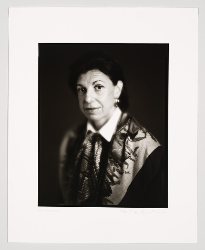 A black and white portrait of a woman with short hair, wearing earrings, a patterned scarf, and a dark blazer. The photograph is framed with a white border that includes a signature and a name at the bottom.
