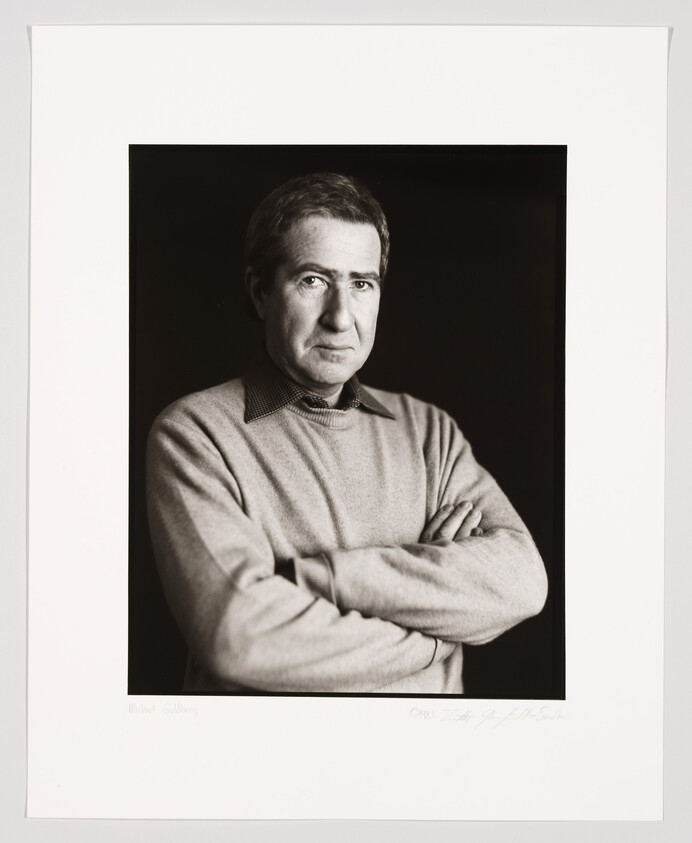 Black and white portrait of a man with crossed arms wearing a collared shirt under a sweater, looking directly at the camera with a serious expression. The photo is framed with a white border containing a signature and text at the bottom.