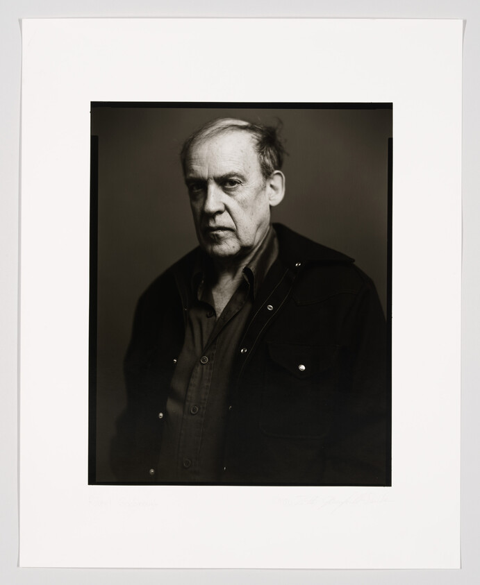 A black and white portrait of an older man with a serious expression, wearing a dark jacket over a buttoned shirt. The photo is framed with a white border, and there is handwriting at the bottom.