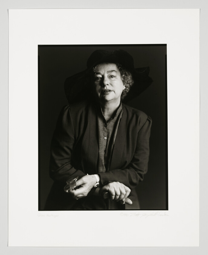 Black and white portrait of a woman with curly hair wearing a large hat and a dark blouse, looking directly at the camera with a calm expression. Her hands are clasped in front of her. The photo is framed with a white border that includes a signature and the name "Grace Hartigan" at the bottom.
