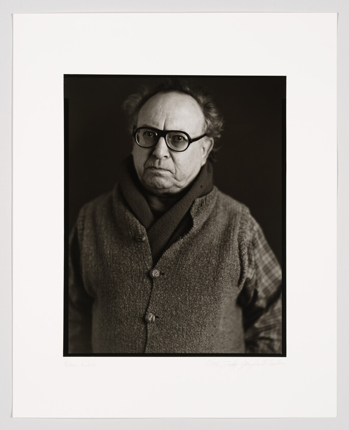 Black and white portrait of a bespectacled man with a serious expression, wearing a scarf and a buttoned cardigan over a plaid shirt. The photograph is framed with a white border that includes a signature and text at the bottom.