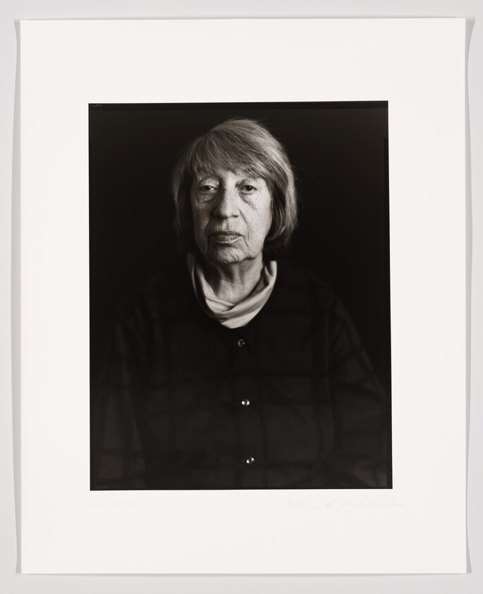 A black and white portrait of an elderly woman with shoulder-length hair, wearing a dark buttoned shirt over a light turtleneck. The photograph is framed with a white border, and there is handwriting at the bottom that appears to be a signature and possibly a name. The background is solid black, emphasizing the subject's face and upper clothing.