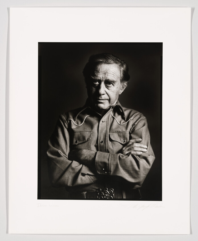 Black and white portrait of an older man with crossed arms, wearing a button-up shirt, with a serious expression. The photo is framed and includes a signature at the bottom.