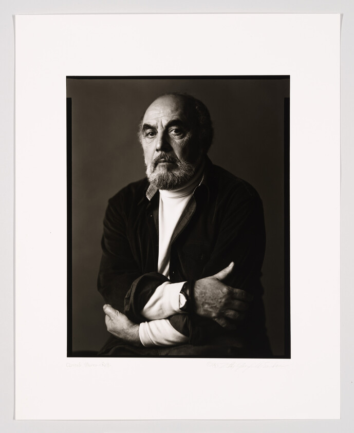 A black and white portrait of a bearded man with a contemplative expression, wearing a dark jacket over a collared shirt, with his arms crossed. The photograph is framed with a white border containing a signature and text at the bottom.