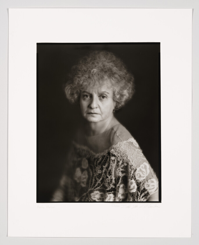 A black and white portrait of an older woman with curly hair, looking directly at the camera with a neutral expression. She is wearing a lace garment. The photograph is framed with a white border, and there is a signature at the bottom.
