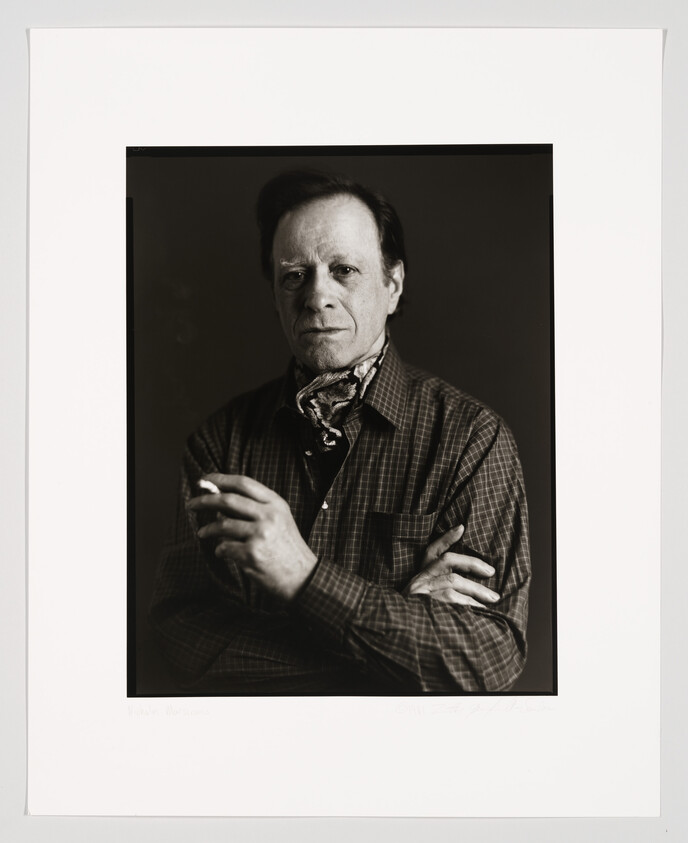 A black and white portrait of a middle-aged man with a serious expression, wearing a plaid shirt and a patterned bandana around his neck. He is looking directly at the camera and his hands are gently clasped in front of him. The photograph is framed with a white border that includes a signature and a name at the bottom.