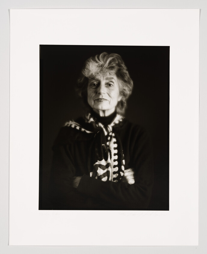 A black and white portrait of an elderly woman with voluminous hair, wearing a dark top with a distinctive light-colored patterned scarf tied at the neck. She is looking directly at the camera with a gentle expression, arms crossed. The photograph is framed with a wide white border.
