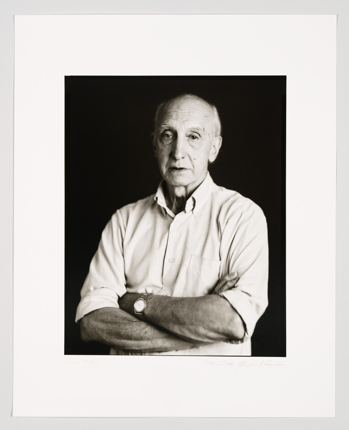 Black and white portrait of an elderly man with a thoughtful expression, wearing a light-colored shirt with his arms crossed. He has a watch on his left wrist and is looking directly at the camera. The photo is framed with a white border that includes a signature and text at the bottom.