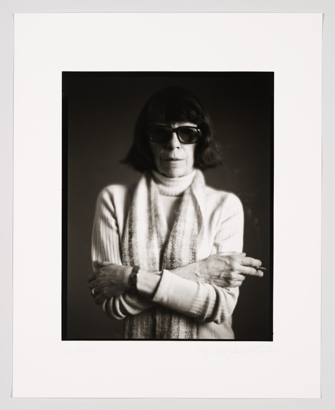 A black and white portrait of a woman wearing sunglasses and a turtleneck sweater, with her arms crossed. The image is framed with a white border, and there is a signature and a name at the bottom.