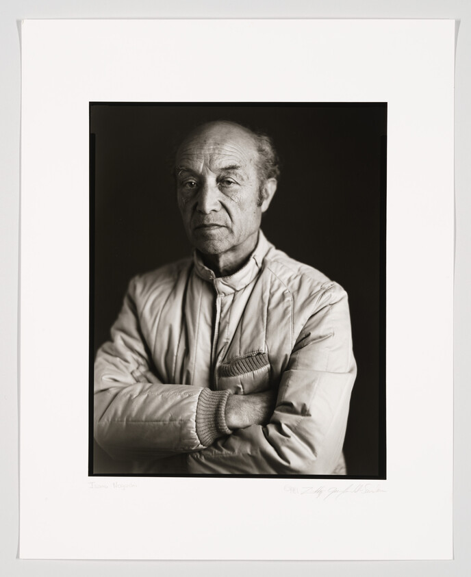 An older man with a serious expression stands with his arms crossed, wearing a light-colored jacket.