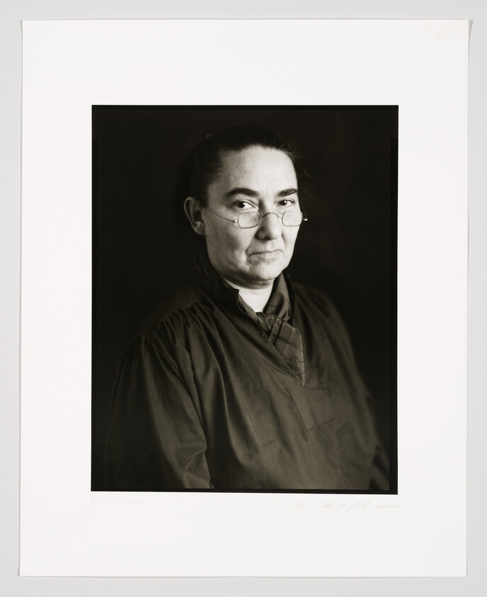 A black and white portrait of a woman with her hair pulled back, wearing round glasses and a dark blouse. The photograph is framed with a white border, and there is handwriting at the bottom.