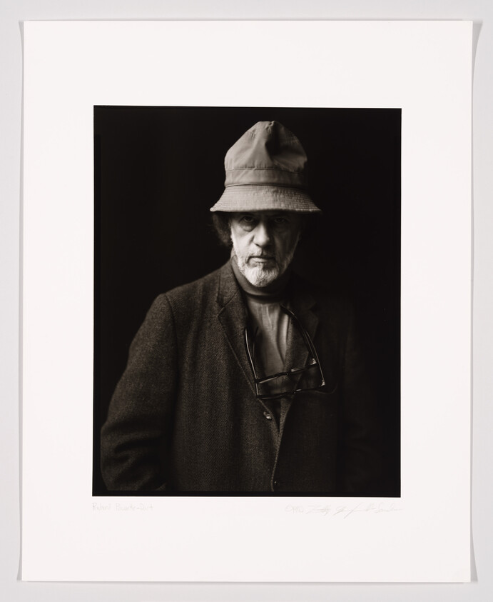 A black and white portrait of an older man with a stern expression, wearing a bucket hat and a coat with glasses hanging from his collar. The photo is signed at the bottom and framed with a white border.