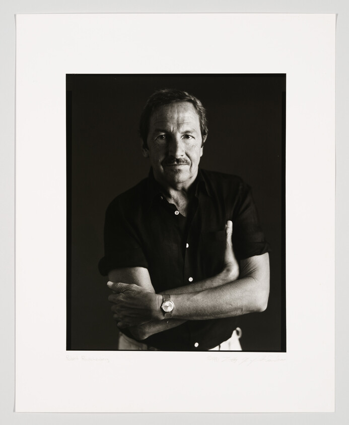 Black and white portrait of a man with a mustache, wearing a black shirt with the sleeves rolled up, and a watch on his left wrist. He is looking directly at the camera with a slight smile and has his arms crossed. The image is framed with a white border that includes a handwritten signature and a name at the bottom.