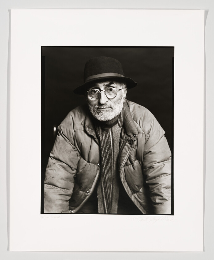 Black and white portrait of a man wearing glasses, a hat, and a jacket, looking directly at the camera with a neutral expression. The photo is framed with a white border, and there is a signature at the bottom left.
