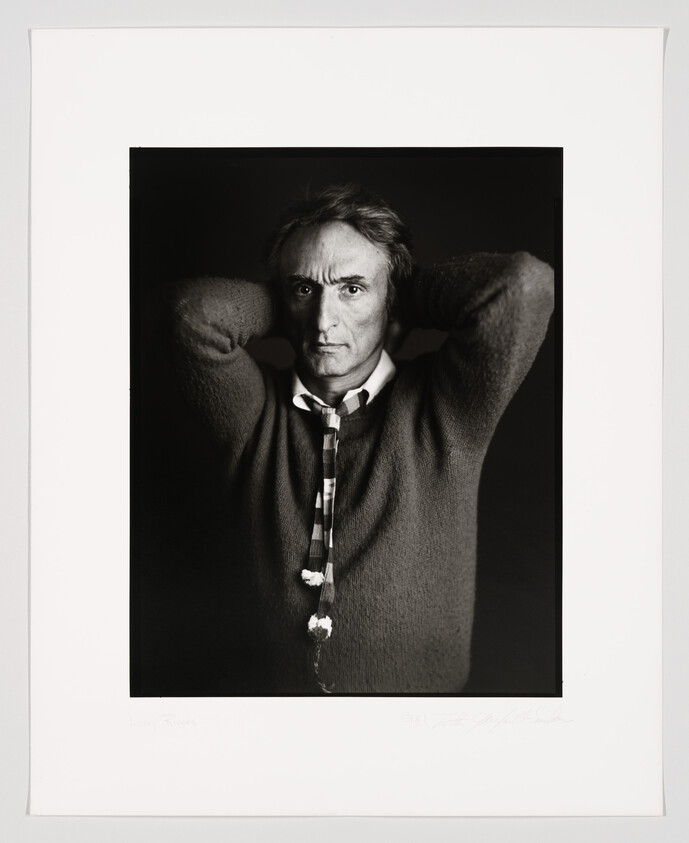 Black and white portrait of a man with intense gaze, hands clasped behind his head, wearing a sweater and a striped tie. The photograph is framed and includes a signature at the bottom.