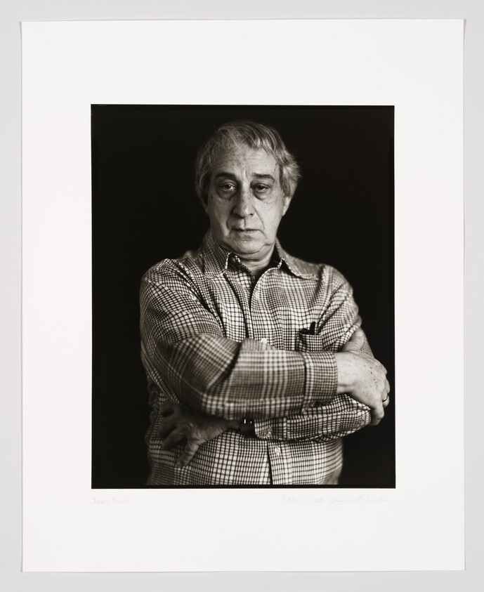 A black and white portrait of an older man with his arms crossed. He is wearing a checkered shirt and has a serious expression on his face. The photograph is framed with a white border, and there is a signature at the bottom.