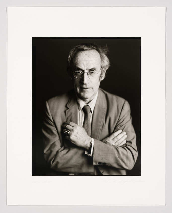 A black and white portrait of a bespectacled middle-aged man with combed-back hair, wearing a suit and tie, with his arms crossed. The image is framed with a white border that includes a signature at the bottom.
