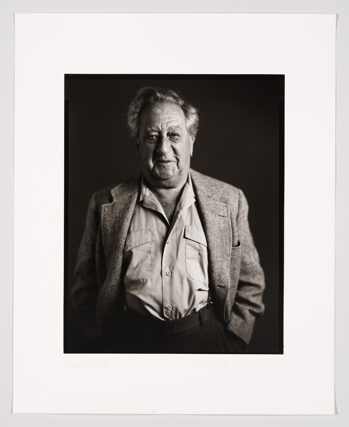 A black and white portrait of an elderly man with a solemn expression, wearing a tweed jacket and an open-collar shirt. The photo is framed and includes a signature and a name written in pencil on the white border below the image.