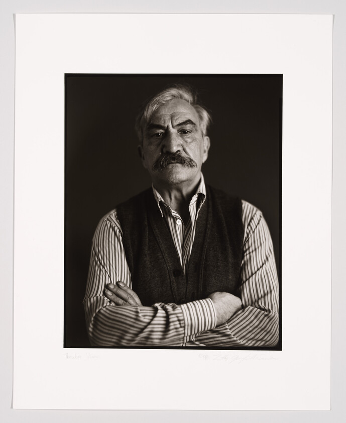 Black and white portrait of an older man with a mustache, wearing a striped shirt and a dark vest, arms crossed, looking directly at the camera. The photo is framed with a white border, and there is handwriting and a signature at the bottom.