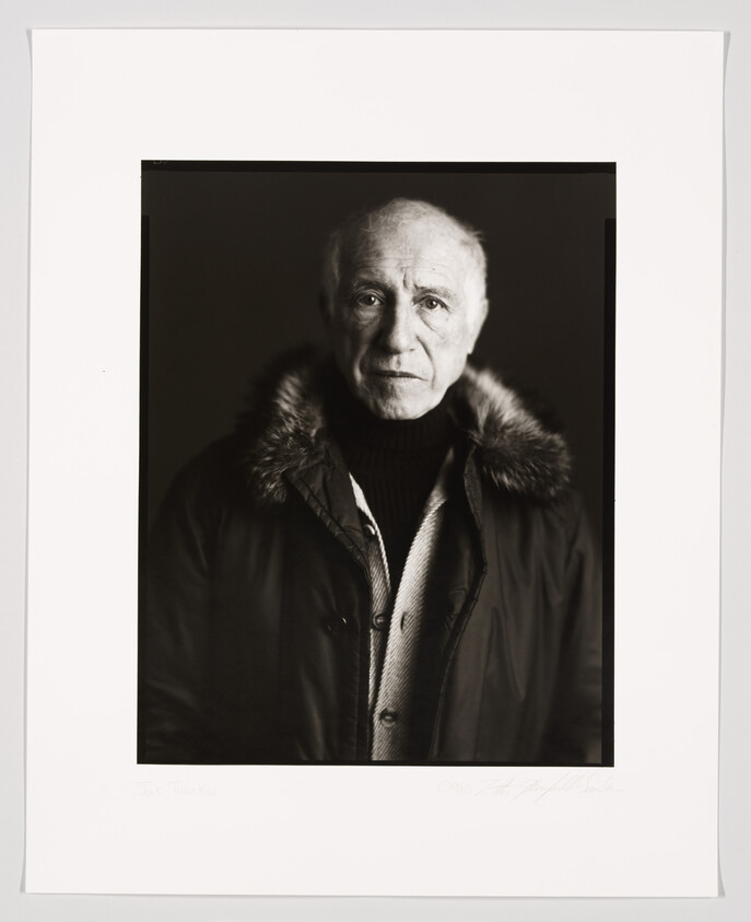 Black and white portrait of an elderly man with a solemn expression, wearing a winter coat with a fur-lined hood and a turtleneck sweater. The photo is framed with a white border containing a signature and text at the bottom.