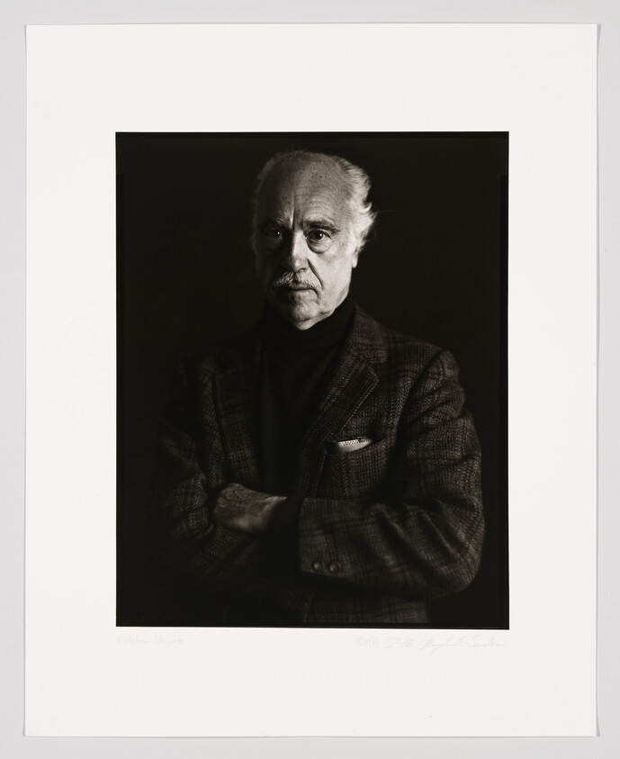 Black and white portrait of an elderly man with a serious expression, wearing a tweed jacket and turtleneck. His arms are crossed, and he has a full head of white hair. The photo is framed with a white border that includes a signature and date at the bottom.