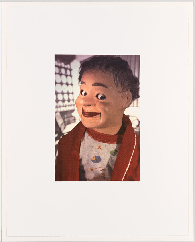 A puppet with a human-like appearance, featuring gray hair and a wide smile, wearing a red cardigan over a shirt with a pattern of flowers and a bird. The background is partially visible, showing a blurred control panel with various buttons and screens.