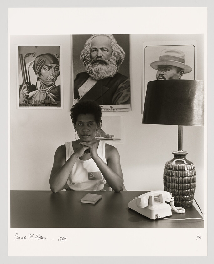 A young black woman sits at a desk with her fists under her chin, political posters and a telephone visible.