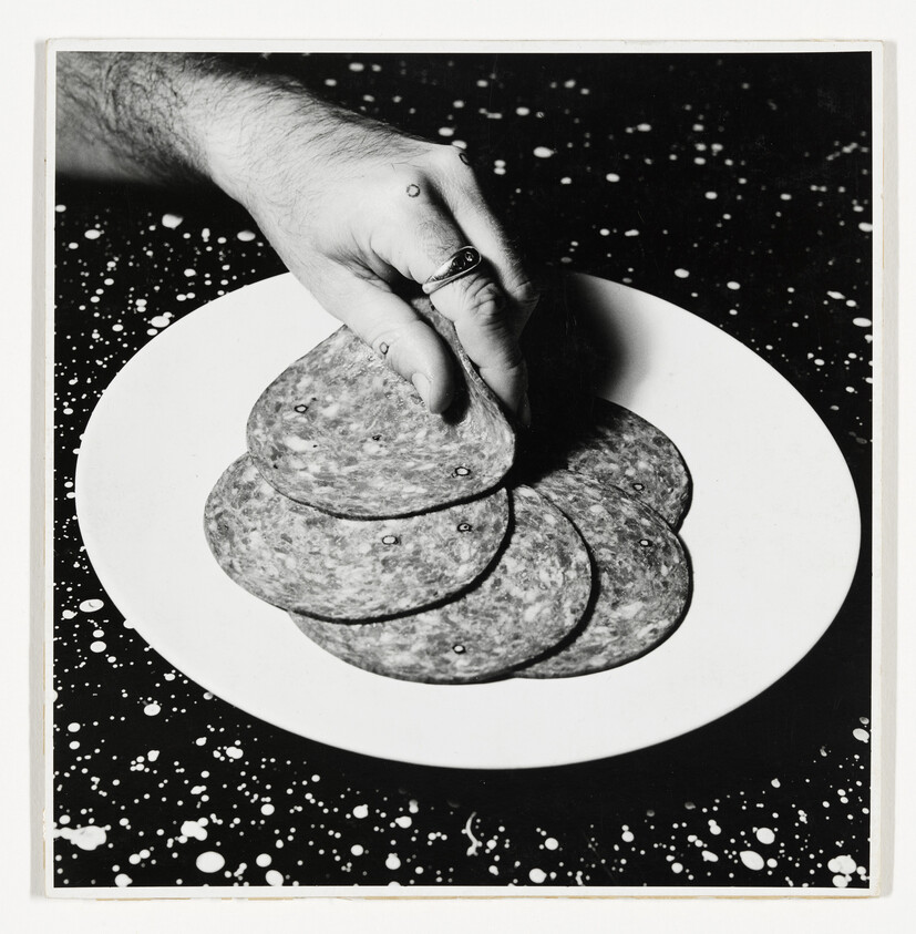 A hand wearing a ring lifts a slice of salami from a stack on a white plate.