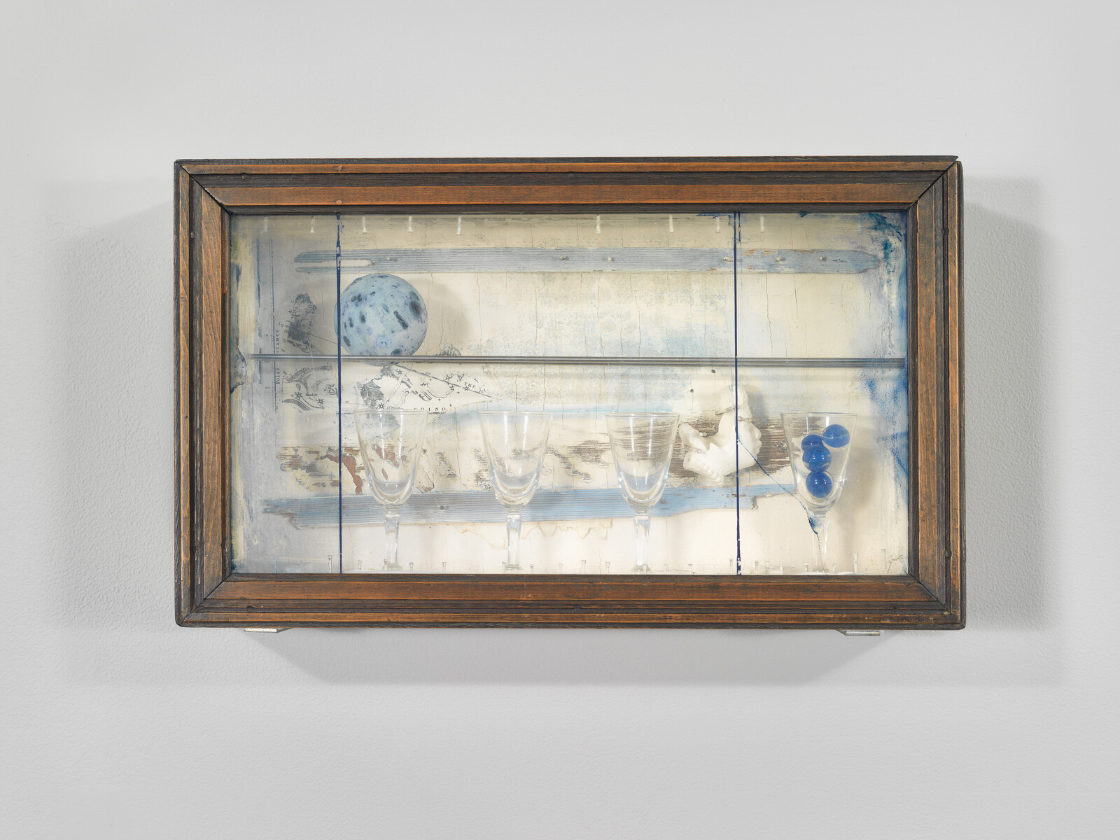 A wooden framed display shows four wine glasses with blue marbles, a speckled ball, and a small white bird.