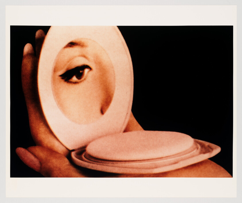 Hand holding a compact mirror showing a woman's eye reflected above a round powder puff.
