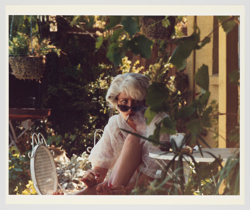 Older woman wearing sunglasses smokes a cigarette while sitting outdoors at a small table.