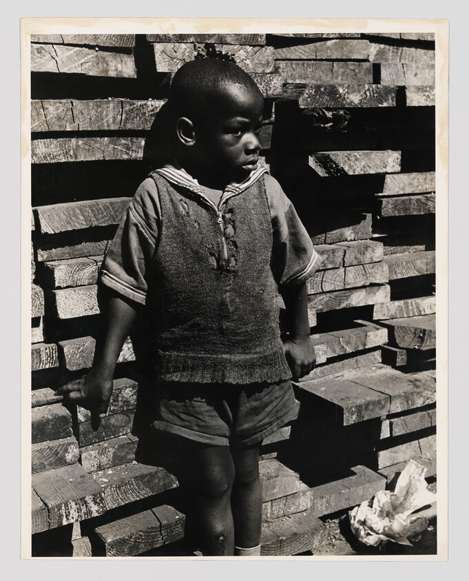 Young boy in worn sweater and shorts standing against a stack of lumber, looking to the side.