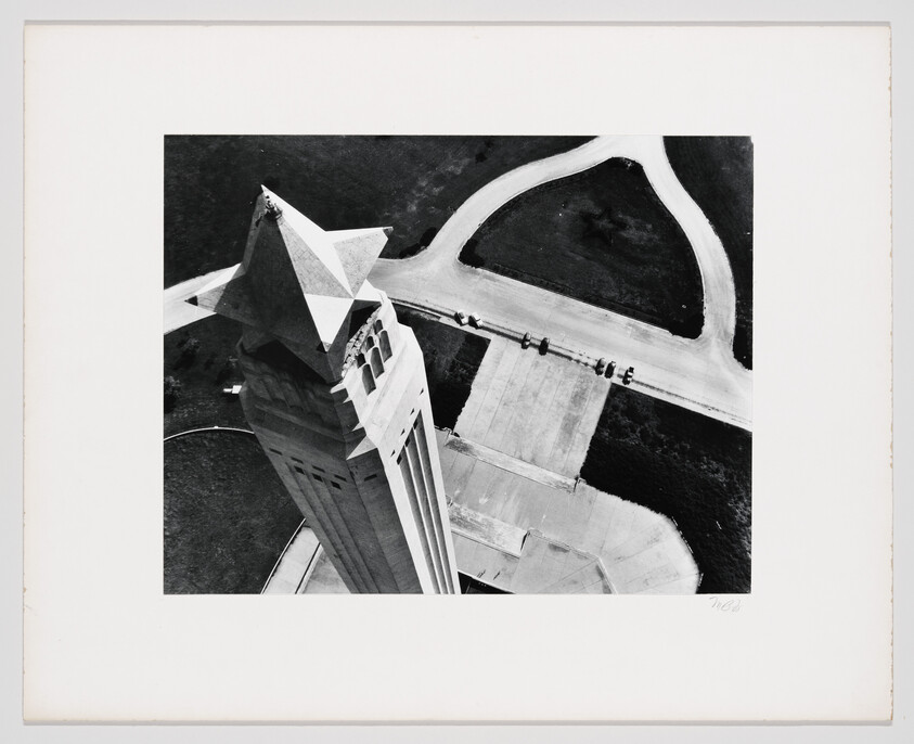 Aerial view looking down a tall stone tower toward a curved road and parked cars.